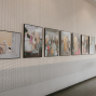 An einer langen, perforierten Galeriewand hängen mehrere gerahmte Fotografien in einer Reihe. Der helle Ausstellungsraum wirkt ruhig, mit Fliesenboden und einer Tür am Ende des Flurs.