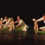 Auf einer dunklen Bühne sitzen drei weiß gekleidete Personen und bewegen weiche, menschenähnliche Puppen. Grüne Folie auf dem Boden und farbiges Licht verleihen der Szene eine experimentelle Theateratmosphäre.