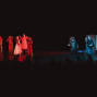 Auf einer dunklen Theaterbühne stehen mehrere kostümierte Darstellerinnen und Darsteller in rotem und blauem Licht verteilt. Im Vordergrund sind die Silhouetten des Publikums zu sehen, während die Szene wie eine dramatische Aufführung oder Tanzsequenz wirkt.