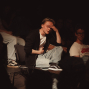 Ein junger Mann sitzt im dunklen Publikum im Scheinwerferlicht auf einem Stuhl und hält sich die Hand an die Stirn. Um ihn herum sitzen weitere Personen im Halbdunkel, wodurch eine konzentrierte, theaterartige Atmosphäre entsteht.