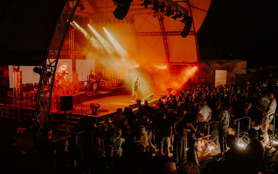 Auf einer überdachten Bühne tritt eine Band vor einem dicht gedrängten Publikum auf, während der Sänger im Zentrum im Lichtkegel steht. Rote und orange Scheinwerfer sowie Nebel erzeugen eine dramatische, energiegeladene Konzertatmosphäre.