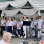 Eine Gruppe von Sängerinnen und Sängern in weißen T-Shirts steht auf einer geschmückten Bühne mit weißen Stoffbahnen. Eine blonde Frau singt ins Mikrofon, während ein Mann mit Gitarre und weitere Musiker neben Keyboard und Mikrofonständern bereitstehen.