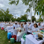 Viele Menschen in weißer Kleidung sitzen an langen, weiß gedeckten Tischen bei einem festlichen Picknick im Park. Zwischen den Bäumen hängen weiße Bänder, während ein bewölkter Himmel über der geselligen Veranstaltung liegt.