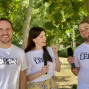 Drei lächelnde Personen stehen in einem grünen Park unter Bäumen und tragen weiße T-Shirts mit der Aufschrift „über LEBEN“. Zwei von ihnen halten Mikrofone in der Hand, während die Frau in der Mitte lachend zu dem Mann rechts schaut.
