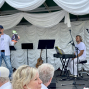 Auf einer Bühne mit weißen Stoffdrapierungen spricht ein Mann mit Mikrofon zum Publikum und hält ein Heft hoch. Rechts sitzt ein Musiker am Keyboard, während mehrere Mikrofonständer und eine Gitarre für eine Aufführung bereitstehen.