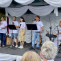 Eine Gruppe von Sängerinnen und Sängern steht auf einer dekorierten Bühne und tritt mit Mikrofonen vor Publikum auf. Rechts begleitet ein Musiker die Darbietung am Keyboard, während weiße Stoffbahnen den Bühnenhintergrund schmücken.