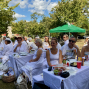 Eine große Gruppe von Menschen in weißer Kleidung sitzt an langen, weiß gedeckten Tischen bei einem Picknick im Park. Auf den Tischen stehen Getränke, Obst und Geschirr, während im Hintergrund Bäume, ein grünes Zelt und ein blauer Himmel mit Wolken zu sehen sind.