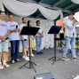 Auf einer Bühne stehen mehrere Menschen in weißen T-Shirts mit Mikrofonen, während ein Mann rechts in ein Mikrofon spricht. Die Bühne ist mit weißen Stoffbahnen dekoriert, und vor der Gruppe stehen Notenständer sowie ein Keyboard im Hintergrund.