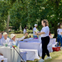 In einem grünen Park sitzen mehrere ältere Menschen in weißer Kleidung an festlich gedeckten Tischen, während eine junge Frau ihnen Flyer oder Unterlagen reicht. Im Hintergrund stehen weitere Personen in weißen T-Shirts zwischen den Bäumen, und die Szene wirkt wie eine sommerliche Gemeinschaftsveranstaltung im Freien.