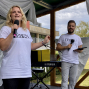 Eine blonde Frau steht auf einer kleinen Außenbühne und spricht mit einem Mikrofon, während sie ein weißes T-Shirt trägt. Neben ihr hält ein Mann ebenfalls ein Mikrofon und ein Klemmbrett; im Hintergrund sind weiße Stoffbahnen, ein Yamaha-Keyboard und ein bewölkter Himmel zu sehen.