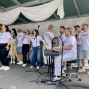 Eine Gruppe von Sängerinnen und Sängern steht auf einer überdachten Bühne und singt mit Mikrofonen, während ein Mann am Keyboard spielt. Alle tragen weiße T-Shirts, und die Bühne ist mit weißen Stoffbahnen dekoriert.