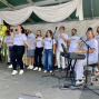 Eine Gruppe von Sängerinnen und Sängern in weißen T-Shirts steht auf einer Bühne und singt fröhlich in Mikrofone. Rechts begleitet ein Musiker die Gruppe am Keyboard, während weiße Stoffbahnen die Bühne dekorieren.