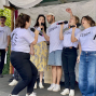 Eine Gruppe von Sängerinnen und Sängern steht auf einer kleinen Bühne im Freien und singt mit Mikrofonen. Alle tragen weiße T-Shirts mit der Aufschrift „über LEBEN“, im Hintergrund sind weiße Stoffbahnen, Bäume und eine Gitarre zu sehen.