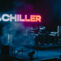 Auf einer dunklen Konzertbühne leuchtet im Hintergrund der große Schriftzug „SCHILLER“ in Blau, Weiß und Rot. Rechts sitzt ein Musiker zwischen Synthesizern, Keyboards und Schlagzeug, umgeben von Nebel und atmosphärischem Bühnenlicht.