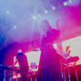 Eine Sängerin steht mit erhobener Hand auf einer Konzertbühne, während Musiker an elektronischen Instrumenten spielen. Bunte pinke und blaue Lichter sowie eine große LED-Wand erzeugen eine atmosphärische Showkulisse.