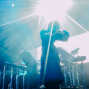 Auf einer Konzertbühne steht eine Sängerin im Gegenlicht, während grelle Scheinwerfer und blauer Nebel die Szene dominieren. Im Hintergrund spielen weitere Musiker an Gitarren, Schlagzeug und elektronischen Instrumenten.