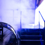 Im Vordergrund steht ein großer Ventilator, der von blau-violettem Licht angestrahlt wird. Im Hintergrund sind unscharf eine Treppe, Geländer und ein Bühnenscheinwerfer in einer atmosphärischen Veranstaltungsszene zu sehen.