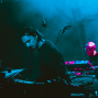 Ein Musiker steht auf einer dunkel beleuchteten Bühne am Mikrofon, umgeben von Schlagzeug und technischem Equipment. Im Hintergrund spielt ein weiterer Musiker an einem Keyboard, während blaues und pinkes Licht die Szene prägen.