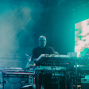 Ein Musiker steht auf einer nebligen Bühne und bedient mehrere Keyboards und Synthesizer. Türkisfarbenes Licht, Bühnenrauch und eine große helle Leinwand im Hintergrund erzeugen eine atmosphärische Konzertstimmung.