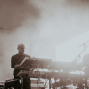 Ein Musiker steht auf einer nebligen Bühne und bedient mehrere Synthesizer und Keyboards. Helles Gegenlicht und Mikrofonständer im Hintergrund erzeugen eine atmosphärische Konzertszene.