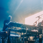 Ein Musiker steht auf einer dunkel beleuchteten Bühne und spielt an mehreren Keyboards und Synthesizern. Blaues Licht, Nebel, Kabel und Mikrofonständer erzeugen eine atmosphärische Konzertszene.