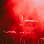 Ein Musiker steht auf einer dunklen Bühne hinter elektronischen Instrumenten und einem Laptop. Rotes Licht und dichter Nebel tauchen die Szene in eine dramatische, fast abstrakte Atmosphäre.