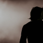 Eine Person ist als dunkle Silhouette vor einem hellen, verschwommenen Hintergrund zu sehen. Links und rechts stehen Mikrofone auf Stativen, was auf eine Bühnen- oder Aufnahmesituation hindeutet.