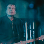 Ein älterer Musiker steht auf einer dunkel beleuchteten Bühne und hält eine elektrische Gitarre. Blaugrünes Licht, Scheinwerfer und ein unscharfer Hintergrund erzeugen eine atmosphärische Konzertstimmung.