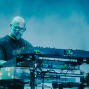 Ein kahlköpfiger Musiker mit Brille steht auf einer blau beleuchteten Bühne und bedient mehrere elektronische Keyboards und Geräte. Um ihn herum sind Kabel, Mischpulte und technisches Equipment zu sehen, während im Hintergrund Nebel oder Projektionen eine atmosphärische Konzertstimmung erzeugen.