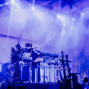 Auf einer Konzertbühne steht ein Musiker zwischen zahlreichen Synthesizern, Kabeln und technischen Geräten, umgeben von dichtem Nebel und blau-violettem Licht. Rechts sind Gitarren und ein weiterer Musiker zu sehen, während im Hintergrund das Publikum im Halbdunkel sitzt.