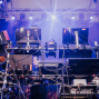 Auf einer Bühne steht ein komplexes elektronisches Musik-Setup mit vielen Monitoren, Kabeln, Halterungen und Steuergeräten. Im Hintergrund leuchten blaue und violette Scheinwerfer durch Nebel, während ein Musiker teilweise hinter der Technik zu sehen ist.