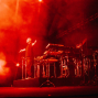Ein Musiker steht mit ausgebreiteten Armen auf einer Bühne, die in intensives rotes Licht und dichten Nebel getaucht ist. Um ihn herum sind Keyboards, Percussion und ein Schlagzeug aufgebaut, während Scheinwerfer die dramatische Konzertatmosphäre verstärken.