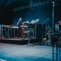 Eine Band steht auf einer dunkel beleuchteten Bühne mit Synthesizern, Gitarren und einem Schlagzeug. Der Sänger hält ein Mikrofon in der Hand, während die Musiker von blauem Bühnenlicht umgeben sind.