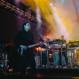 Ein Musiker spielt auf einer Bühne E-Gitarre, während im Hintergrund ein weiterer Künstler an mehreren Keyboards und elektronischen Geräten steht. Bunte Scheinwerfer, Nebel und große Lichtprojektionen erzeugen eine dramatische Konzertatmosphäre.