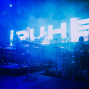 Eine Band spielt auf einer dunkel beleuchteten Bühne, die in intensives blaues Licht und Nebel getaucht ist. Im Hintergrund leuchtet eine große LED-Wand mit riesigen weißen Buchstaben, während Schlagzeug, Mikrofone und elektronische Instrumente zu sehen sind.