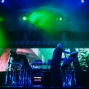 Ein Musiker steht auf einer dunklen Bühne vor mehreren Synthesizern und elektronischen Geräten. Grüne und blaue Scheinwerfer, Nebel und große Videoprojektionen erzeugen eine intensive Konzertatmosphäre.
