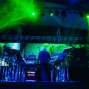Auf einer Konzertbühne steht ein Musiker von hinten vor Synthesizern und elektronischem Equipment, während grüne Laserstrahlen und blaues Licht die Szene erhellen. Im Hintergrund sind große LED-Projektionen mit tropischen Blattmustern sowie Schlagzeug und Bühnenkonstruktionen zu sehen.