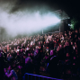 Eine große Menschenmenge sitzt und steht bei einer nächtlichen Open-Air-Veranstaltung und applaudiert. Über dem Publikum liegen dichter Nebel und grelle Scheinwerferstrahlen, die die Szene dramatisch beleuchten.
