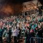 Eine große Menschenmenge steht nachts auf einer Tribüne und hält leuchtende Smartphones in die Höhe. Über den Zuschauern strahlen Bühnenlichter, während Rauch und Lichtreflexe eine Konzert- oder Veranstaltungsatmosphäre erzeugen.