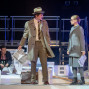 Auf einer Theaterbühne stehen ein Mann im Mantel und Hut sowie ein Mädchen mit Brille einander gegenüber, beide halten Zeitungen in der Hand. Im Hintergrund sitzt eine weitere Person zwischen weißen Bühnenrequisiten, während blaues Licht und Metallgerüste die Szene beleuchten.
