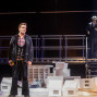 Auf einer Theaterbühne steht ein Mann im Vordergrund in dunkler Kleidung und kariertem Hemd zwischen weißen Modellhäusern und Requisiten. Im Hintergrund befindet sich ein weiterer Mann im Anzug mit Hut auf einer erhöhten Plattform neben einer beleuchteten Treppe.
