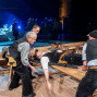 Auf einer Theaterbühne tragen mehrere Männer einen bewusstlos wirkenden Mann auf einer Trage über einen Holztisch. Im Hintergrund stehen weitere Darsteller in blauem Bühnenlicht zwischen Kulissen und Requisiten.