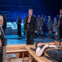 Auf einer Theaterbühne stehen mehrere Darsteller in Arbeitskleidung unter blauem Licht, während ein Mann in der Mitte ernsthaft nach vorn blickt. Im Vordergrund liegt ein Mann auf einem Holztisch, daneben steht ein weiterer Darsteller mit Werkzeugtasche und Headset.