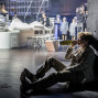 Auf einer Theaterbühne sitzt eine Person im Anzug im Schatten an einer schwarzen Wand und trinkt aus einer Flasche. Im Hintergrund sind ein helles Bühnenbild, Stühle und mehrere weitere Darsteller im Scheinwerferlicht zu sehen.