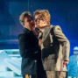 Auf einer blau beleuchteten Theaterbühne stehen zwei Schauspieler eng beieinander, wobei einer den anderen am Revers festhält. Im Hintergrund sind Bühnenaufbauten und weitere Darsteller in gedämpftem Licht zu sehen.