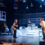 Auf einer Theaterbühne stehen mehrere Schauspieler in einer modern-industriellen Kulisse mit Metallgerüsten, hängenden Lampen und weißen Modellhäusern. Zwei Männer stehen sich im Vordergrund gegenüber, während weitere Personen auf Podesten und im Hintergrund die Szene beobachten.