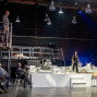 Auf einer großen Theaterbühne stehen und sitzen mehrere Schauspieler auf verschiedenen Ebenen zwischen Metallgerüsten, weißen Bühnenmodellen und hängenden Lampen. Rechts agiert ein Mann im weißen Unterhemd energisch, während andere Figuren ihn beobachten und im Hintergrund Musiker zu sehen sind.