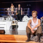 Auf einer Theaterbühne sitzt ein junger Mann in Gummistiefeln und weißem Unterhemd nachdenklich am Bühnenrand. Im Hintergrund stehen weitere Darsteller auf einer erhöhten Kulisse mit Geländern und weißen Modellhäusern, während blaues Bühnenlicht die Szene beleuchtet.