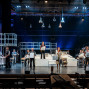 Auf einer großen Theaterbühne stehen zahlreiche Darstellerinnen und Darsteller mit weißen Modellhäusern und Bauteilen in den Händen. Die Szene ist modern und technisch inszeniert, mit Gerüsten, Treppen, hängenden Lampen und einer Musikergruppe im Hintergrund.