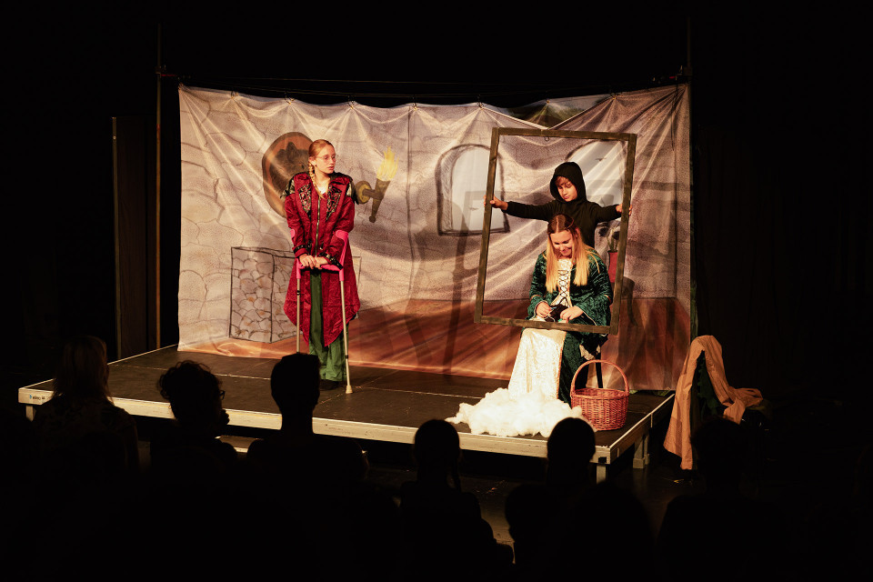 Auf einer kleinen Theaterbühne stehen drei kostümierte Darsteller vor einer bemalten Kulisse, während im Vordergrund die Silhouetten des Publikums zu sehen sind. Eine Person in rotem Gewand stützt sich auf Krücken, während rechts eine sitzende Frau in einem Rahmen von einer schwarz gekleideten Figur gehalten wird.